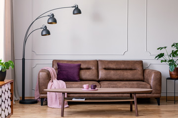 Purple pillow and pastel pink blanket placed on leather couch in real photo of bright sitting room interior with metal lamp, coffee table with tea cups and wainscoting on wall