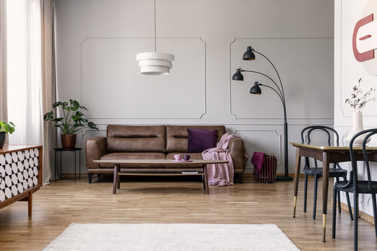 Real photo of light grey sitting room interior with brown couch with blanket and violet pillow, coffee table with two tea cups, metal lamp and molding on wall