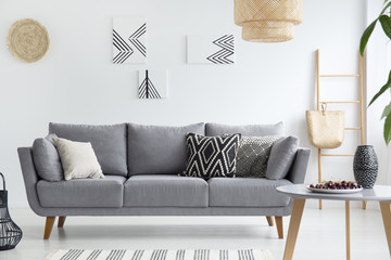 Pillows on grey sofa in white living room interior with posters, lamp and wooden table. Real photo