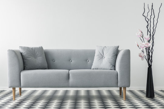 Flowers next to grey sofa with cushions in minimal living room interior with patterned floor. Real photo