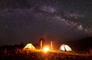 Night camping. Bright campfire burning between two hikers, man and woman standing opposite each other in front of shining tents under amazing dark starry sky with Milky way on distant hills background