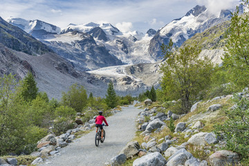 Fototapeta na wymiar Senior woman, riding her e-mountain bike on the trail to the Morteratsch glacier near Pontresina , Engadin, Switzerland,Alps. In the background the famous mountains of Piz palu, Bellavista, Bernina