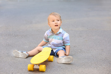 Cute little boy with skateboard sitting on ground outdoors