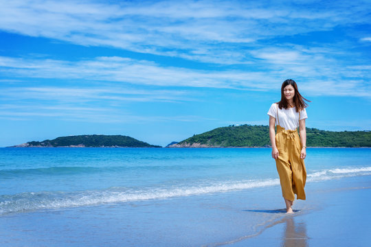 Summer lifestyle image of happy stunning woman walking on the beach of tropical island. Smiling and enjoying life in paradise. Sea background. Put on long brown pants and white shirt.