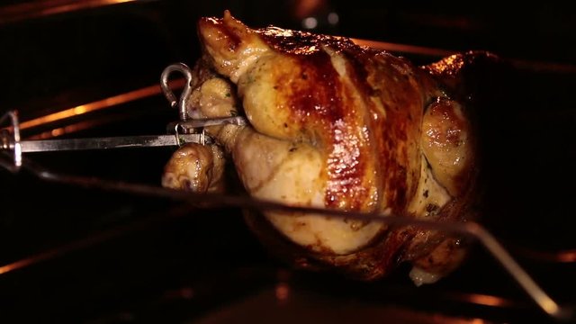 Cooking roasted whole chicken on the rotisserie spit in grilled oven. Browned chicken rotates during broiling under a grill element. Inside view.