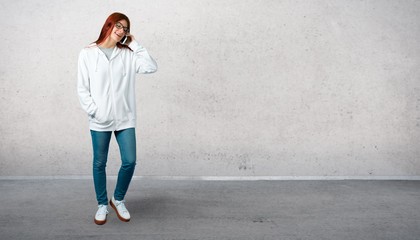 Young redhead girl in an urban white sweatshirt with glasses keeping a conversation with the mobile phone with someone and standing on a vintage gray wall