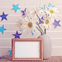 White chamomiles are in a gray vase. Nearby there is an empty wooden frame. On a white wooden background is a paper garland of blue stars.