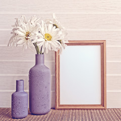 White chamomiles are in a gray vase. The vase is covered with drops of water. Nearby there is an empty wooden frame. White wooden background.