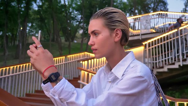 Young modern woman is standing on bridge, takes photo of sunset on smartphone, urban concept, ladder with lights on background
