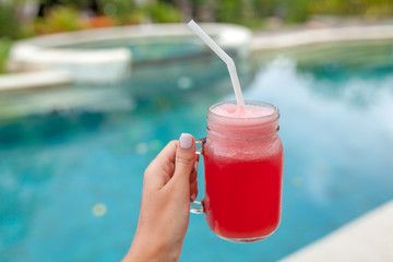 Iced fruit shake in jar in woman hand against pool. Summer and tropical mood. Cold blended drink, watermelon fruit smoothie.
