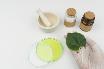 herbal medicine research concept. hand holding an organic green leaf, a mortar and pestle, two bottles and watch glass with green liquid on the white table in laboratory.