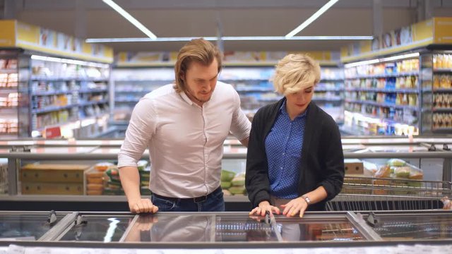 Cute caucasian adult couple choose frozen foods from the supermarket refrigerator. Family shopping at a big trading centre in a grocery department.