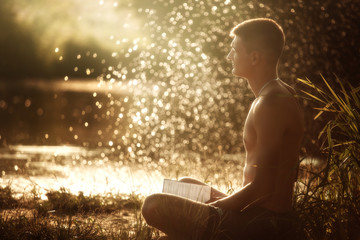 a young man with a naked torso in black shorts sitting on the river bank with a book and medetiruet...