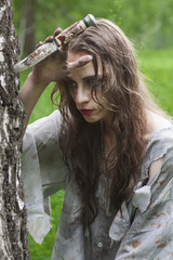Beautiful young dirty mad and manic looking girl wearing torn clothes and smeared with mud and dried blood holds her hand to the knife stuck into tree in the forest