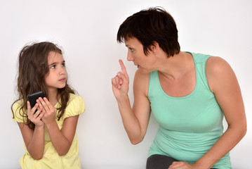 Angry and nervous mother with index finger up scolding her scared daughter with smartphone. Family...