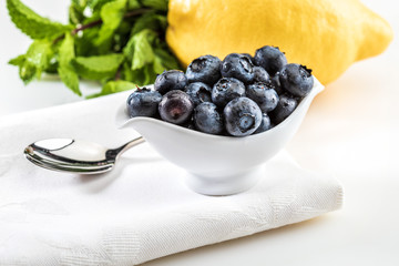 Fresh and raw blueberries in delicate porcelain bowl on white napkin and spoon. Includes brown sugar and lemon. On white background.