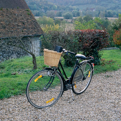 Fototapeta na wymiar Old bicycle with wicker basket on a gravel road in French countryside. Quaint European country scene with shingled house, bicycle and gravel path.