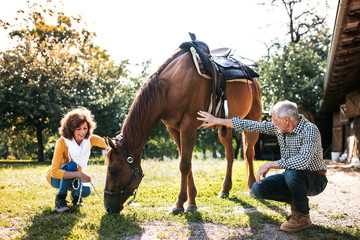 A senior couple crouching and a horse grazing by a stable.