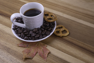 Roasted Coffee beans around coffee espresso mug and two pretzels with fall autumn red maple leaf on wood surface. Rustic coffee during fall - autumn season concept.