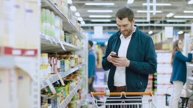 At the Supermarket: Handsome Man Uses Smartphone and Walks Through the Canned Goods Shelf. He's Pushing Shopping Cart through Canned Goods Section.