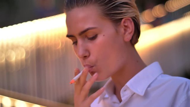 Modern woman is standing and smoking cigarette, watching in smartphone in evening, blurred lights on background