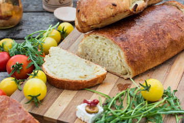 Fresh bread on wooden ground. Yellow tomato and red tomatoes with arugula. Feta cheese with olives and sundried tomatoes. Picnic, dinner outdoor. Still life of food.