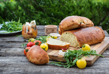 Fresh bread on wooden ground. Yellow tomato and red tomatoes with arugula. Feta cheese with olives and sundried tomatoes. Picnic, dinner outdoor. Still life of food.