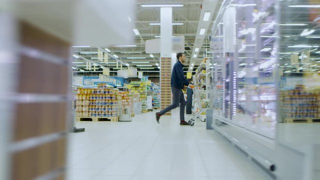 At the Supermarket: Man in a Hurry Pushes Shopping Cart full of Items, He's Walking Through Different Section of the Big Bright Mall. Following / Moving Side view Footage.