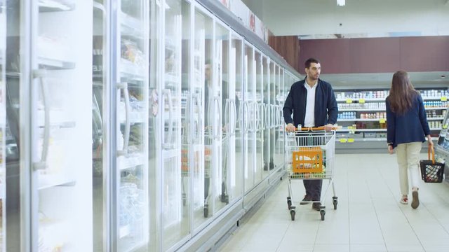At the Supermarket: Handsome Man Pushes Shopping Card and Browses for Products in the Frozen Goods Section. Man Looks into Glass Door Fridge, Looking for Dairy Products. Other Customer Shopping.