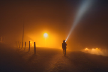 One human silhouette with flashlight in foggy countryside street at night in the orange light of a street lamp © evgenydrablenkov