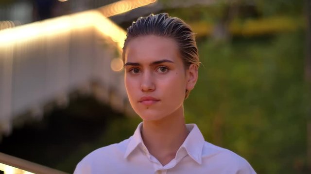 Calm modern woman is watching at camera in evening, blurred lights on background
