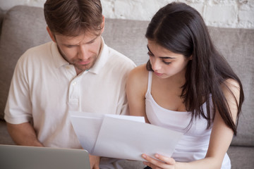 Serious young spouses sitting with laptop and documents, analyzing paperwork and house utility bills, thoughtful couple consider papers managing finances and expenses, checking mail at home