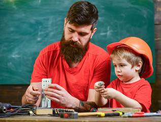 Father with beard and little son in classroom teaching to use tools, chalkboard on background. Boy, child in protective helmet makes by hand, repairing, does crafts with dad. Home education concept.