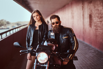 Portrait of an attractive couple - brutal bearded biker in black leather jacket with sunglasses sitting on a motorcycle and his young sensual brunette girlfriend on a footway under a bridge, looking