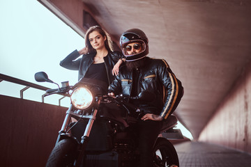 Naklejka na ściany i meble Portrait of an attractive couple - brutal bearded biker in helmet and sunglasses dressed in a black leather jacket sitting on a motorcycle and his young sensual brunette girlfriend on a footway under