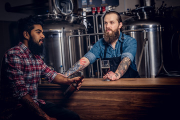 Two bearded interracial friends drink craft beer in a brewery. Tattooed hipster worker male in an apron drinks beer with his friend in the brewery factory.