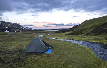 small green tent standing on green grass on creek banks campsite Strutur near road f210, snow patched hills, midnight pink sunset sky, Iceland