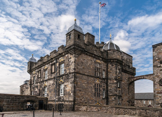 Edinburgh, Scotland, UK - June 14, 2012: Brown gray stone Royal Palace with golden decorations and gate to Crown Square under blue and white sky. Half Moon Battery up front. Brittish flag.