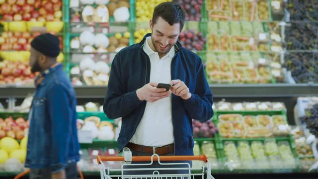 At the Supermarket: Handsome Man Uses Smartphone while Standing in the Fresh Produce Section of the Store. Man Immersed in Internet Surfing on His Mobile Phone In the Background Colorful Fruits