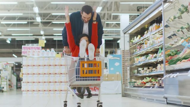 At the Supermarket: Man Pushes Shopping Cart with Woman Sitting in it, Happy Couple Has Fun Racing in a Trolley through the Fresh Produce Section of the Store. 