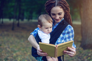 Happy mom with a baby in a sling reading a book in the Park. Children's education in family life