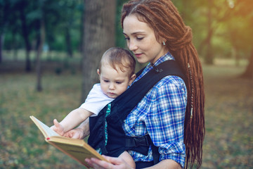 Happy mom with a baby in a sling reading a book in the Park. Children's education in family life