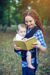 Happy mom with a baby in ergo backpack reading a fairy tale in the Park. Children's education in family life