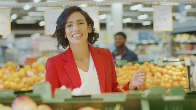 At the Supermarket: Portrait of the Beautiful Smiling Woman Choosing Products In the Fresh Produce Aisle and Places them into Shopping Basket. In the Background Colorful Fruits