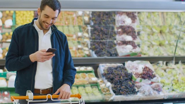 At the Supermarket: Handsome Man Uses Smartphone while Standing in the Fresh Produce Section of the Store. Man Immersed in Internet Surfing on His Mobile Phone In the Background Colorful Fruits. 