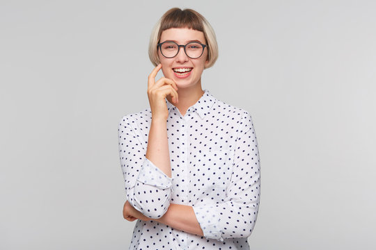 Portrait of happy beautiful blonde young woman wears polka dot shirt and glasses looks joyful, keeps hands folded and laughing isolated over white background