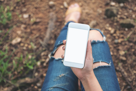 Top view mockup image of woman holding white mobile phone with blank desktop screen while sitting on the ground outdoor