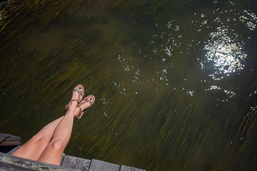 Young woman wearing sandals sitting on a wooden bridge, hanging her legs over the water. female...
