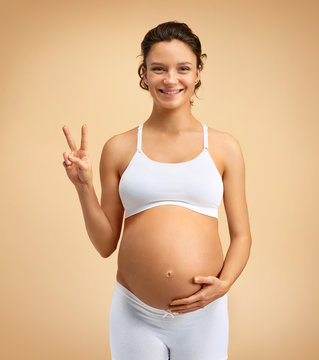 Smiling pregnant woman making two fingers gesture on beige background. Pregnancy, maternity, preparation and expectation concept