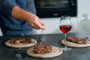 Man putting herbs and spices on the grilled well done steaks. Dinner with red wine.